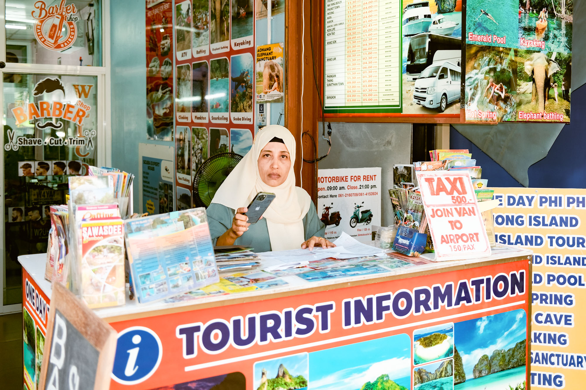 Eine Frau mit einem Smartphone hinter einem Touristenschalter, umgeben von bunten Plakaten und Reiseprospekten.